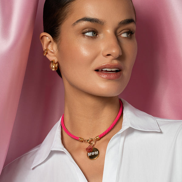 modella indossa un girocollo colore rosa, un charm ed una medaglietta che riporta il testo MOM con cuore centrale. Indossa anche un orecchino ed un earcuff